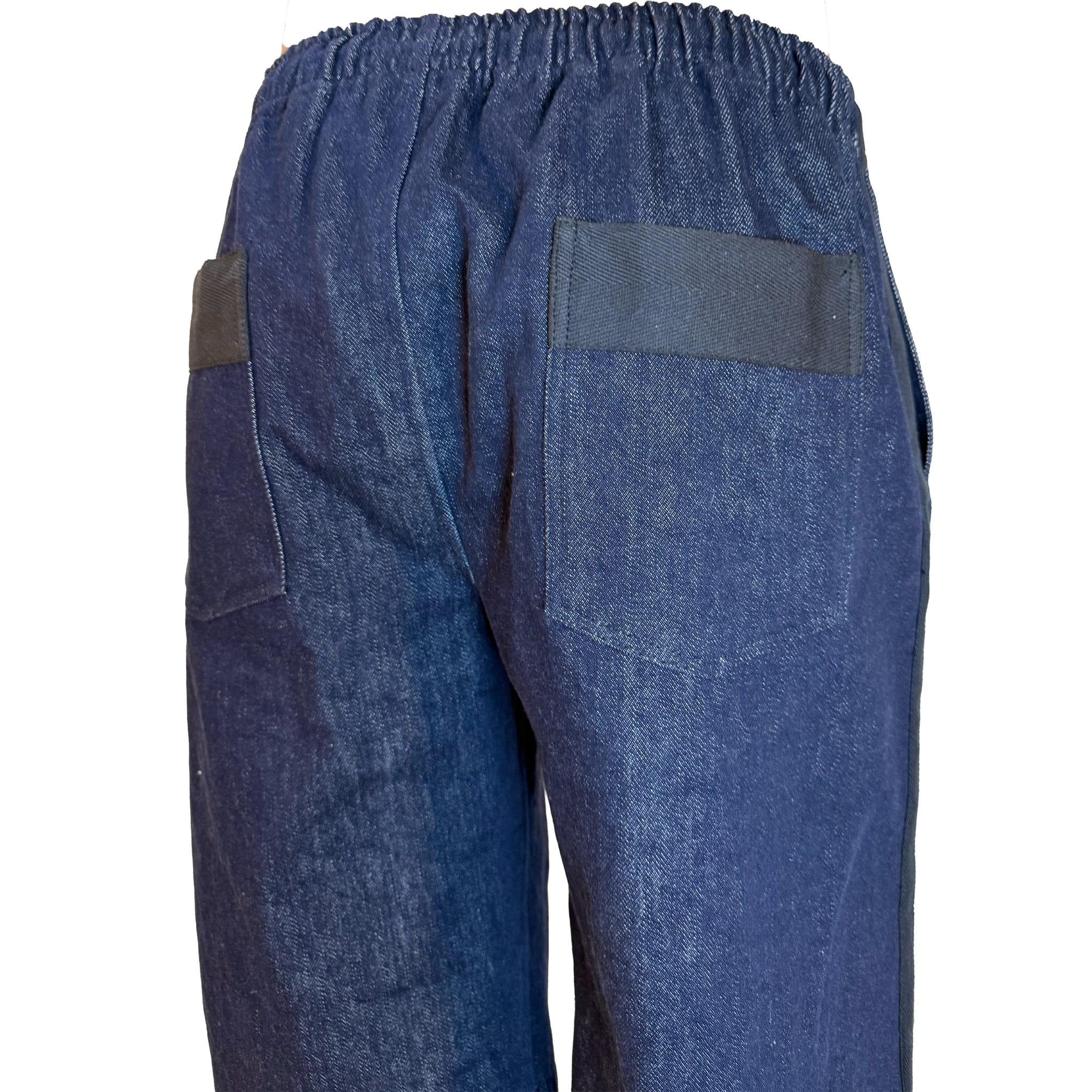Close-up back view of dark blue denim work pants with an elastic waistband, a large back patch pocket, and a dark brown leather patch on the side pocket, shown against a plain white background.
