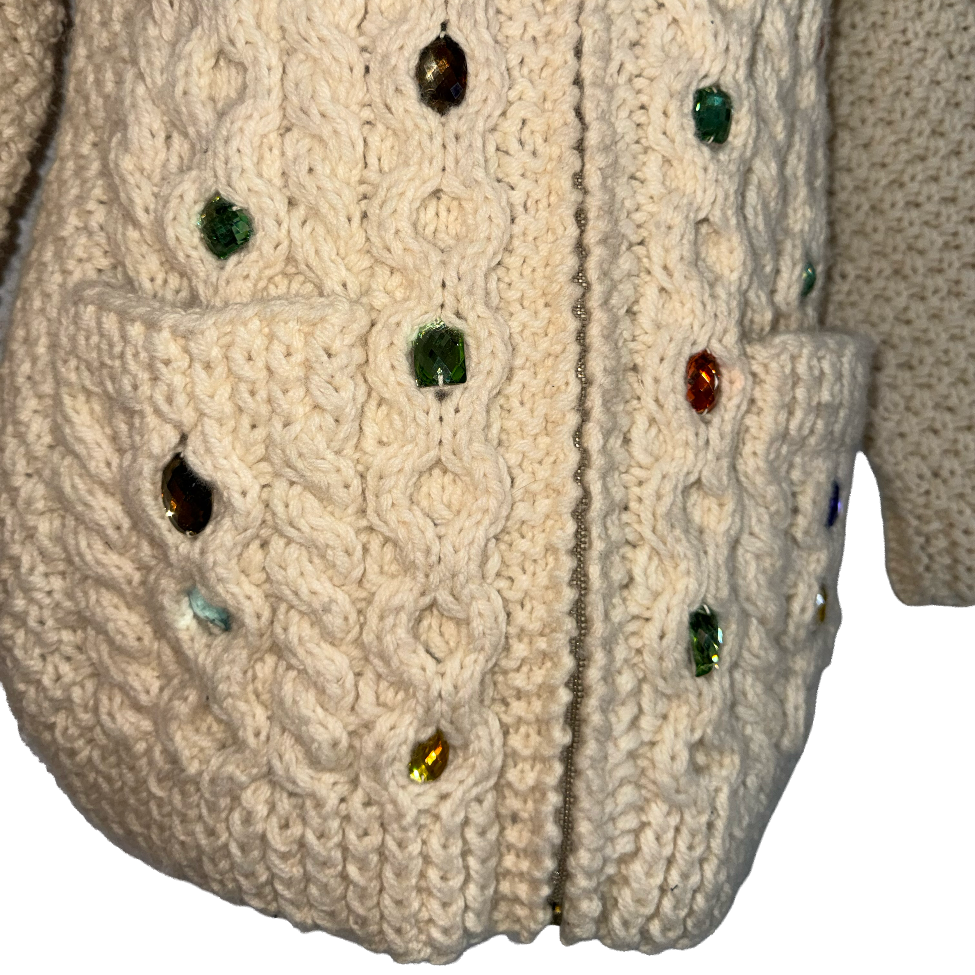 Close-up of a cream cable-knit zip-up cardigan with pockets and colorful faceted rhinestone embellishments, shown against a white background.