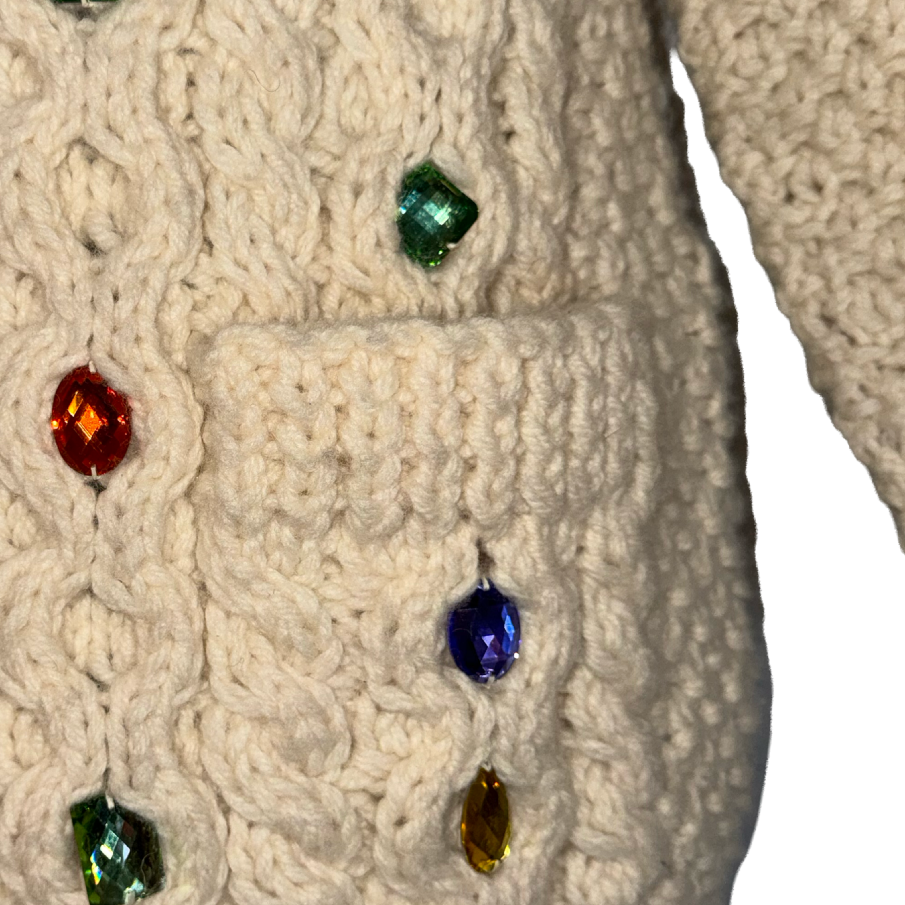 Close-up of a cream-colored cable-knit sweater with a front pocket, decorated with colorful faceted rhinestones in green, blue, red, and gold.