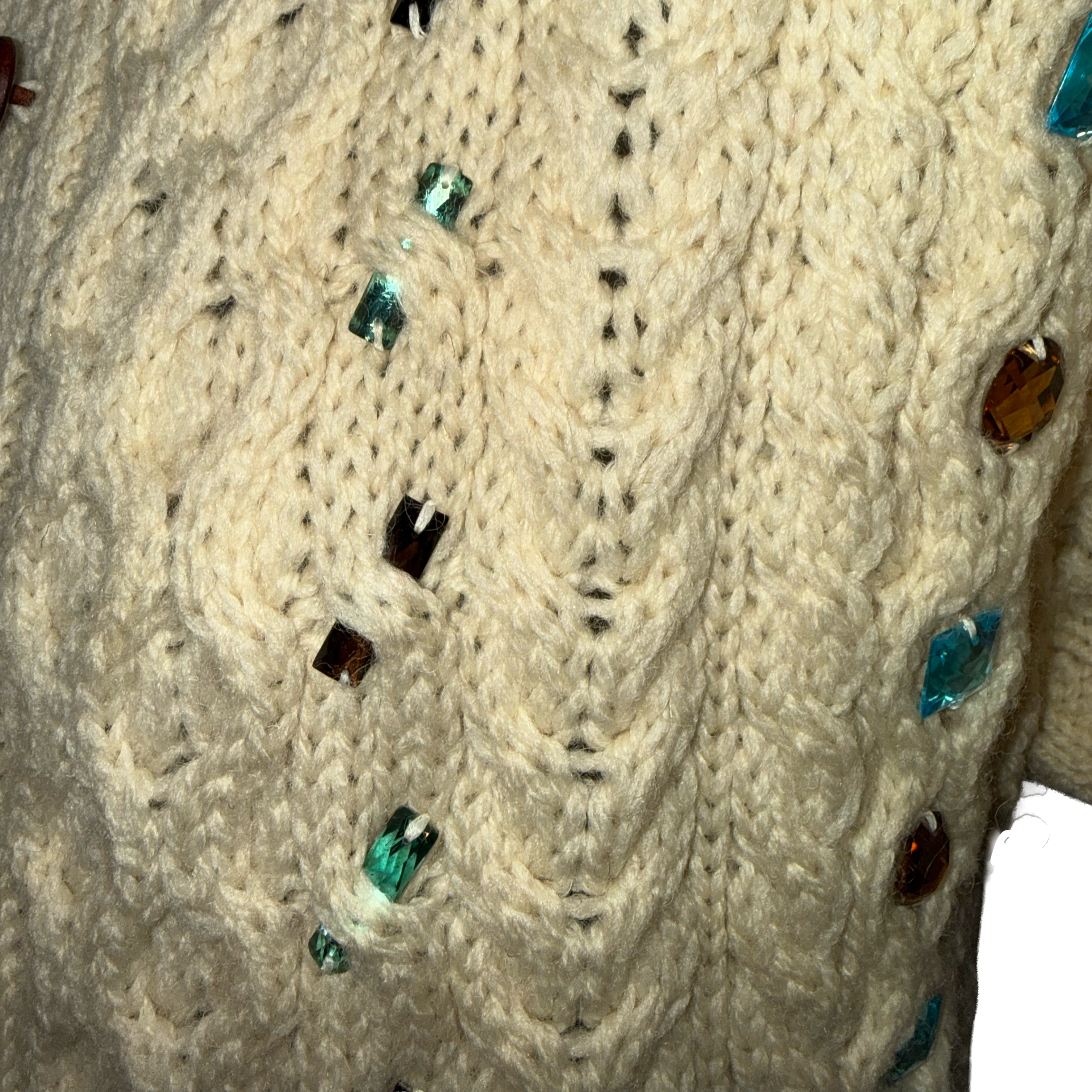 Close-up of a cream cable-knit sweater with scattered faceted rhinestone embellishments in teal, brown, and dark amber.