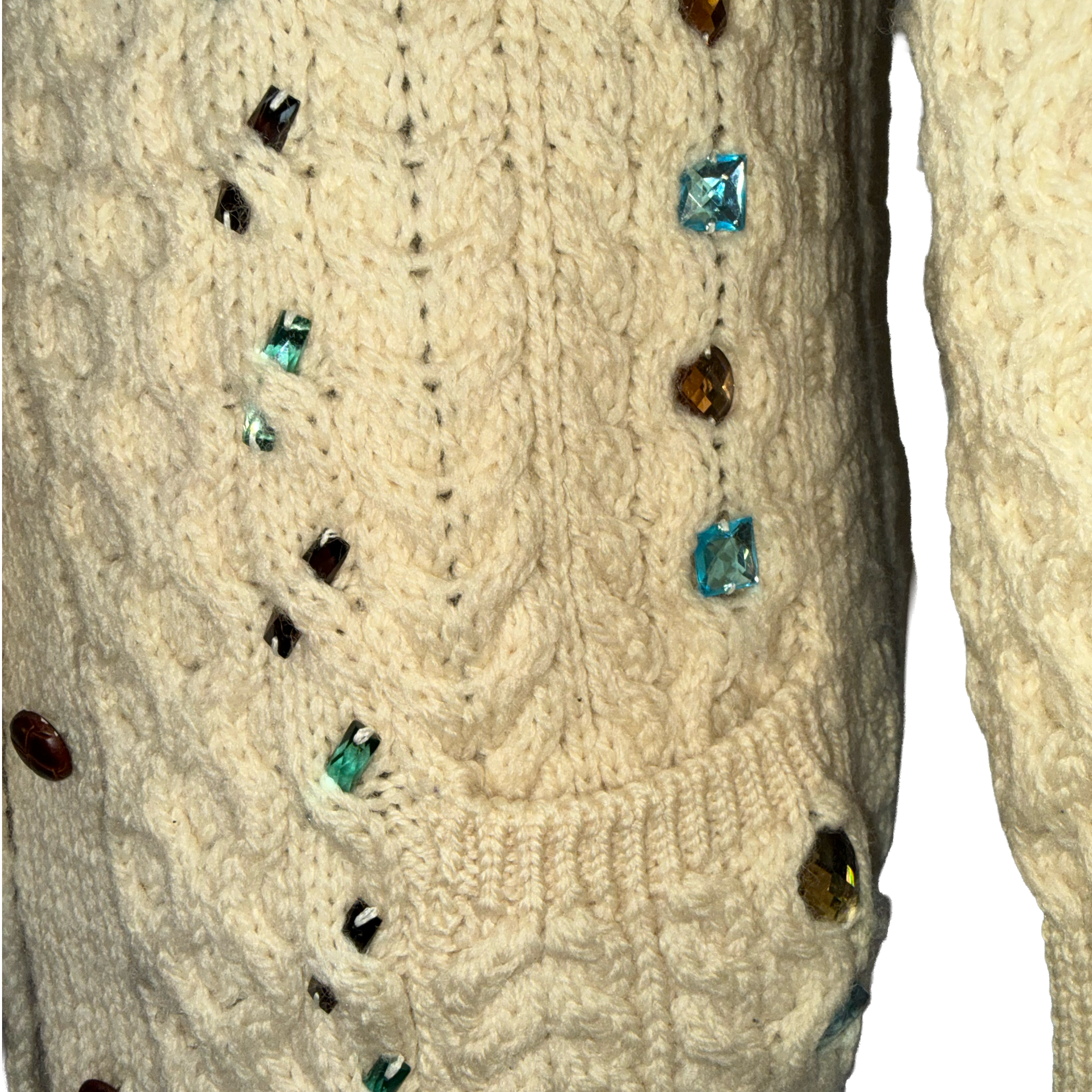 Close-up of a cream cable-knit sweater with decorative faceted gemstones in blue, green, brown, and black scattered across the front, shown against a plain white background.