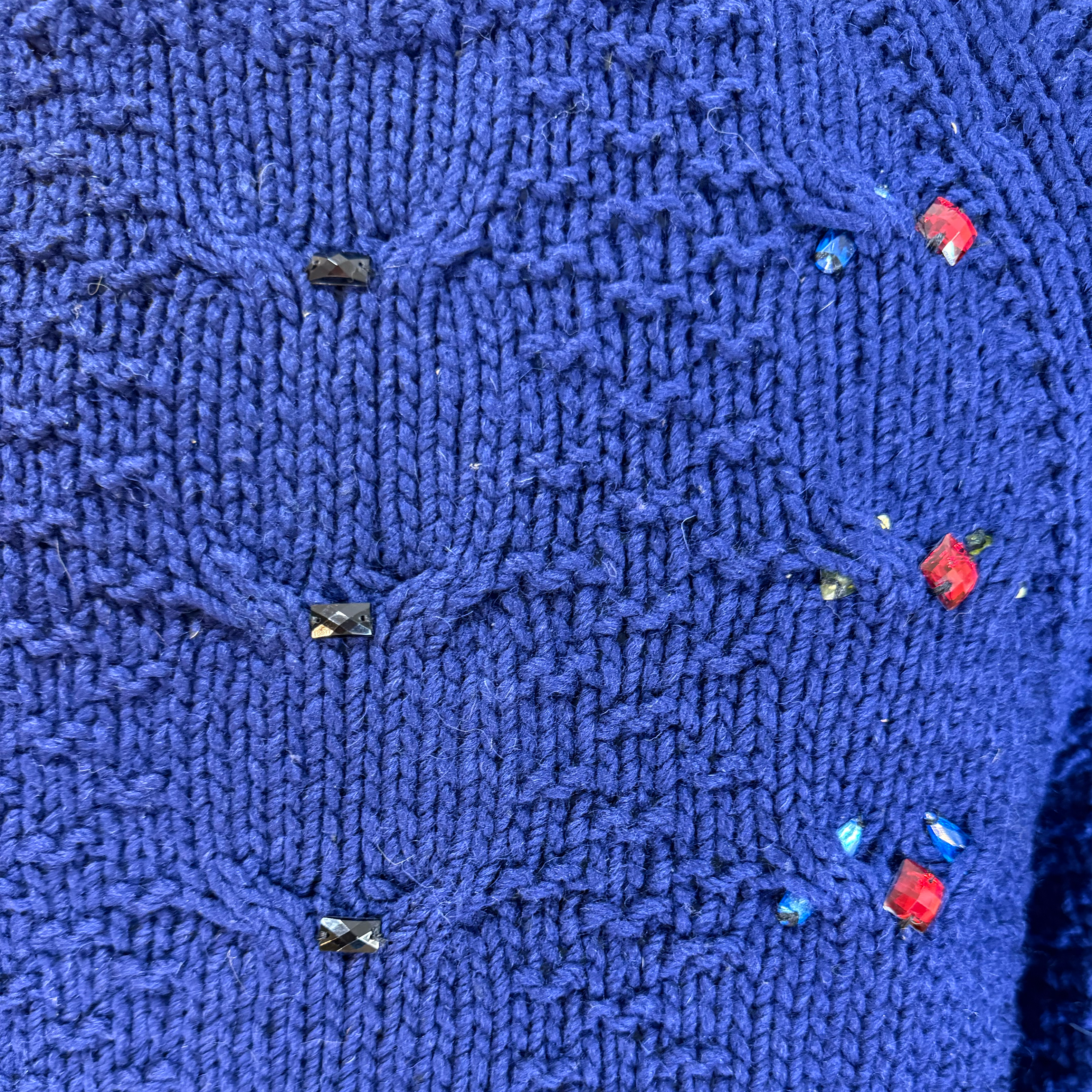 Close-up of a textured royal blue knitted sweater featuring a cable knit pattern and embellished with small rhinestones in red, blue, and black.