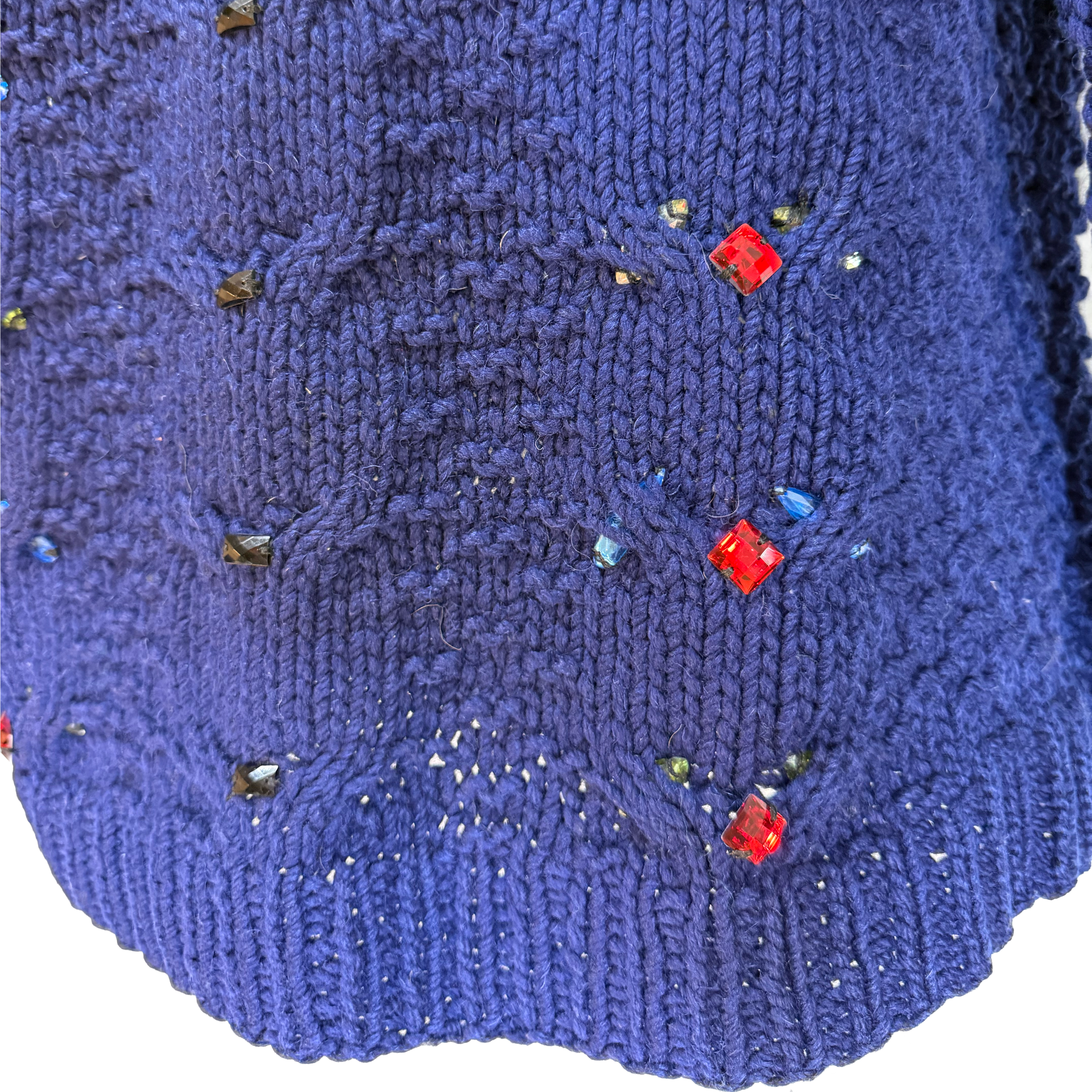 A close-up of a blue knitted garment, likely a sweater or scarf, featuring textured cable and rib patterns with small scattered rhinestone embellishments in red, blue, and green.